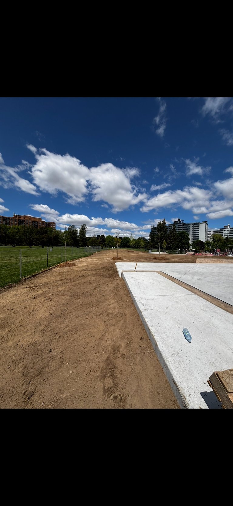 Construction site with concrete foundation in urban park under blue sky with white clouds and surrounding apartment buildings