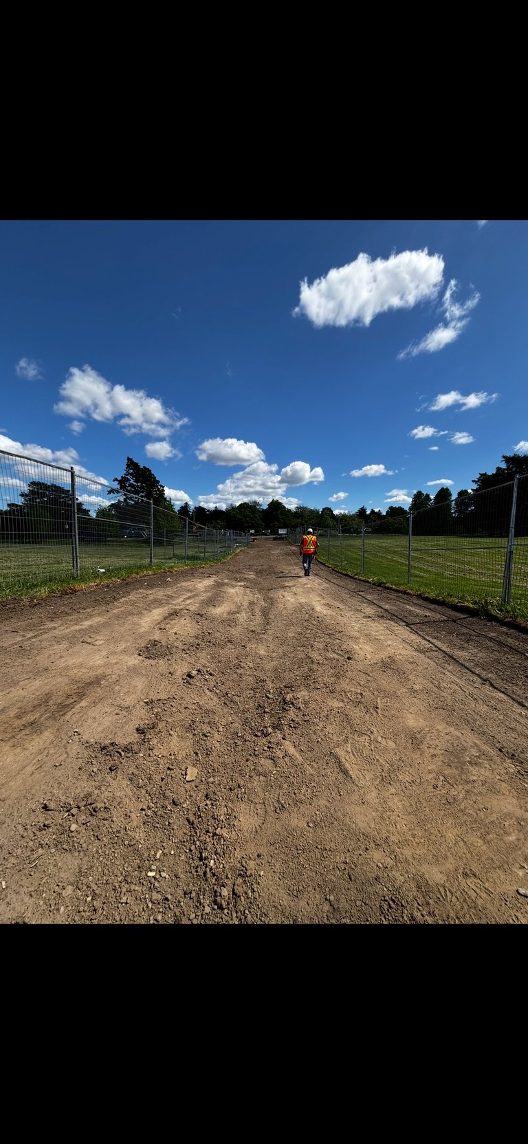 Person in orange vest walking on dirt driveway between metal fences with green pastures and blue sky with white clouds