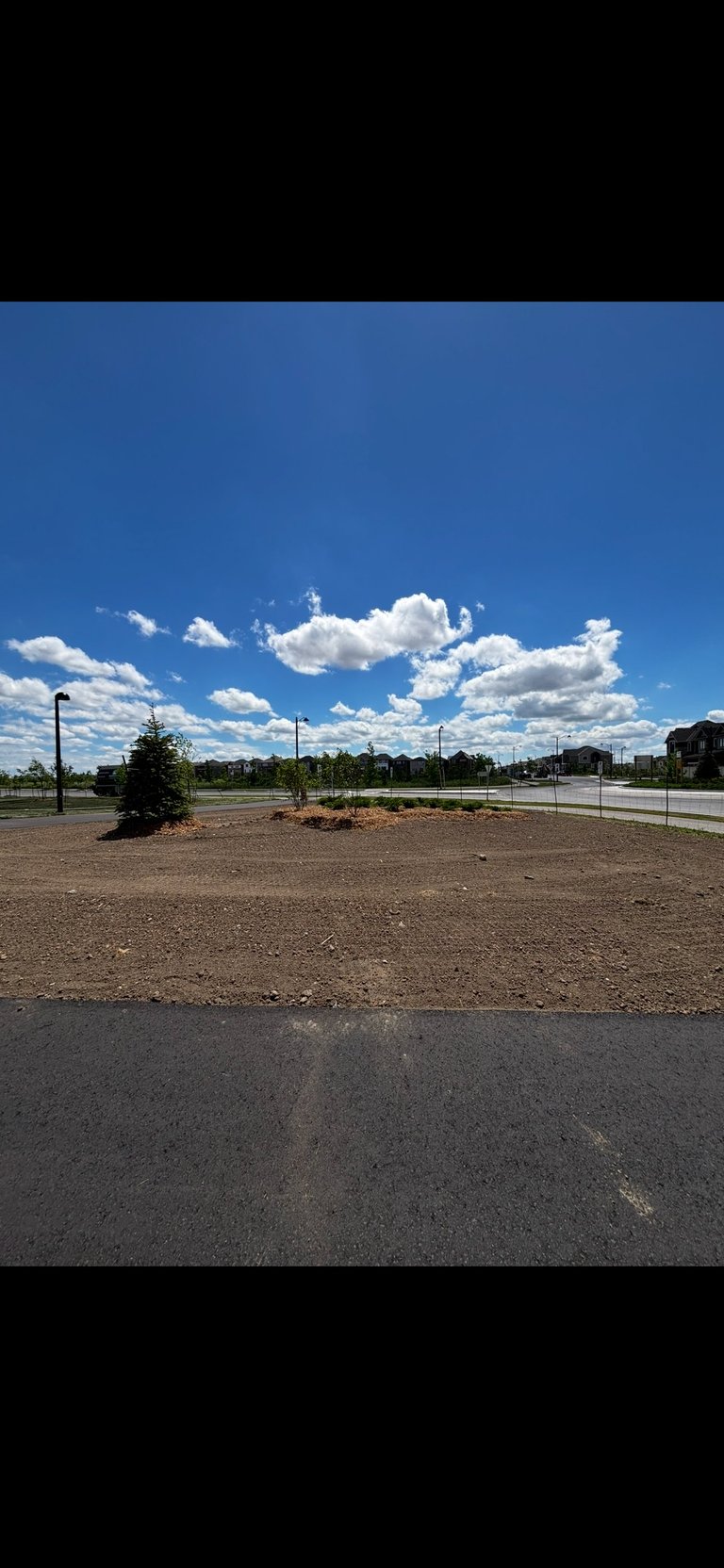 Empty parking lot with dirt and gravel under blue sky with white cumulus clouds and distant trees and buildings
