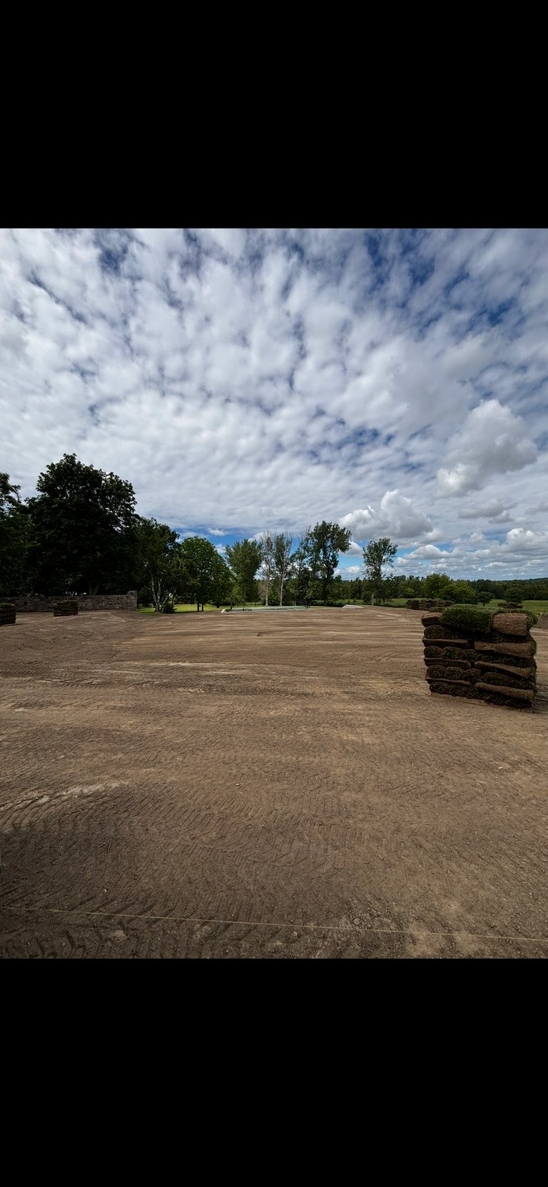 Open dirt arena with stacked hay bales, trees, and dramatic cloudy sky with blue patches visible in background