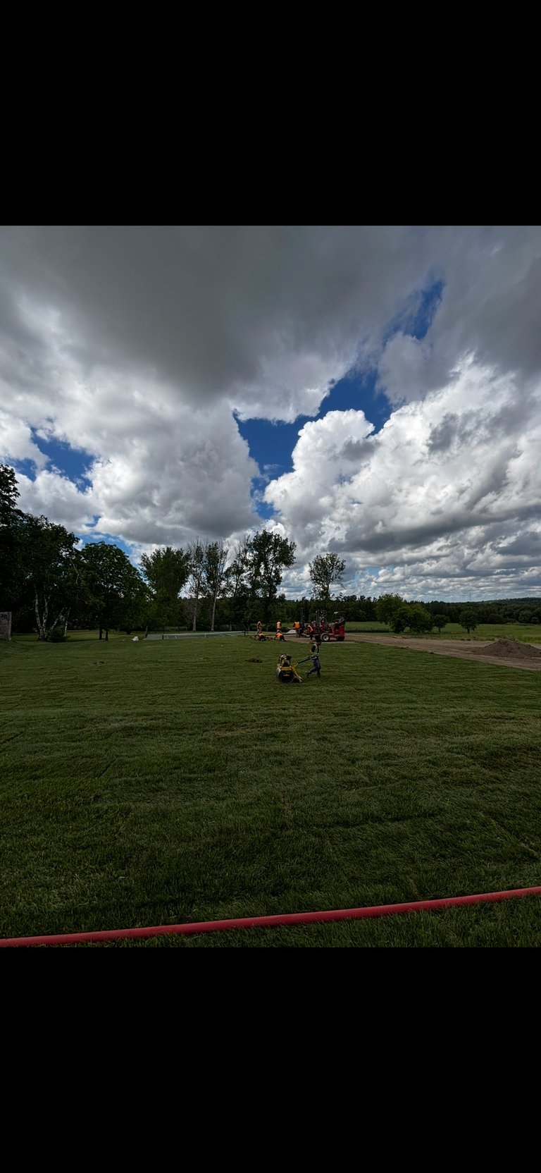 Open park field with sparse trees, people scattered about, dramatic cloudy sky with blue patches, red rope in foreground