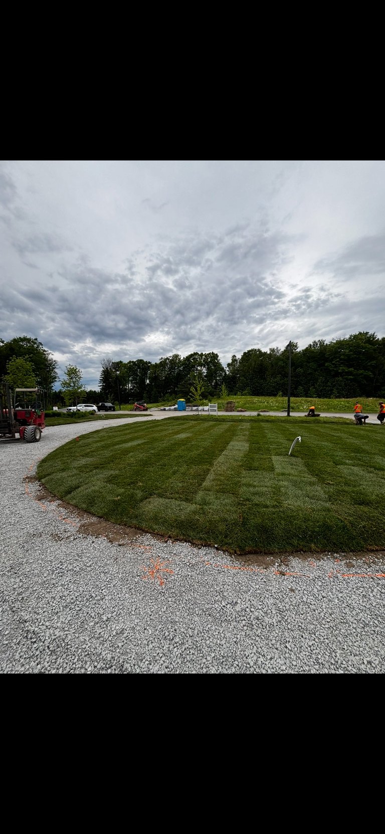 Landscaped lawn with diagonal mowing pattern under stormy sky, surrounded by gravel, trees, and farm equipment