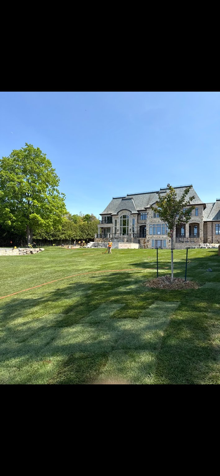Expansive manicured lawn with large luxury stone home featuring turrets and blue shutters, mature trees, and clear blue sky
