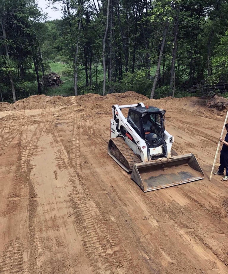 White skid steer loader with front bucket on a dirt construction site surrounded by forest trees